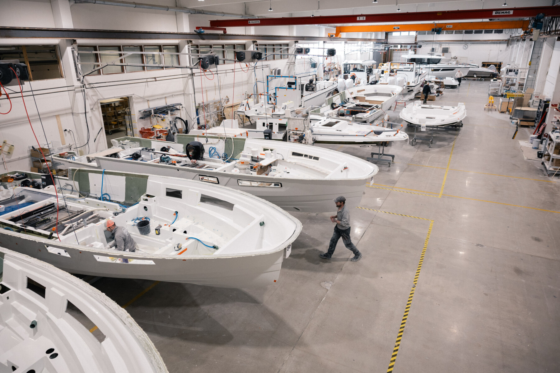 Arbeiter montieren Quarken-Boote in einer großen Fabrikhalle, in der mehrere weiße Rümpfe auf der Produktionsfläche aufgereiht sind.