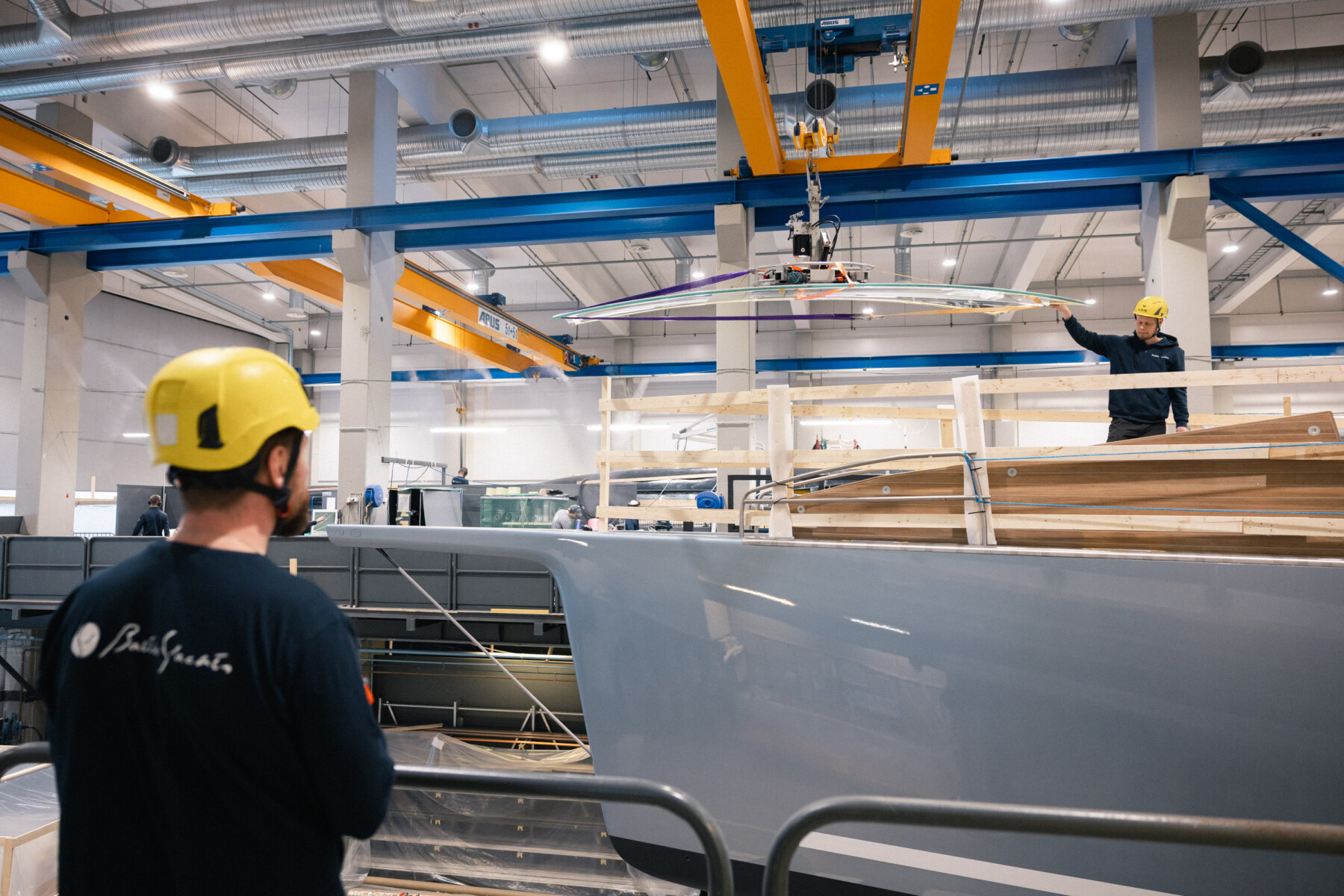 Arbeiter mit Schutzhelmen montieren eine große Luxusyacht in der Produktionshalle von Baltic Yachts.