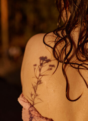 Wet strands of hair fall across a person’s bare shoulder as the shadow of a small wildflower is cast onto their skin in warm evening light.