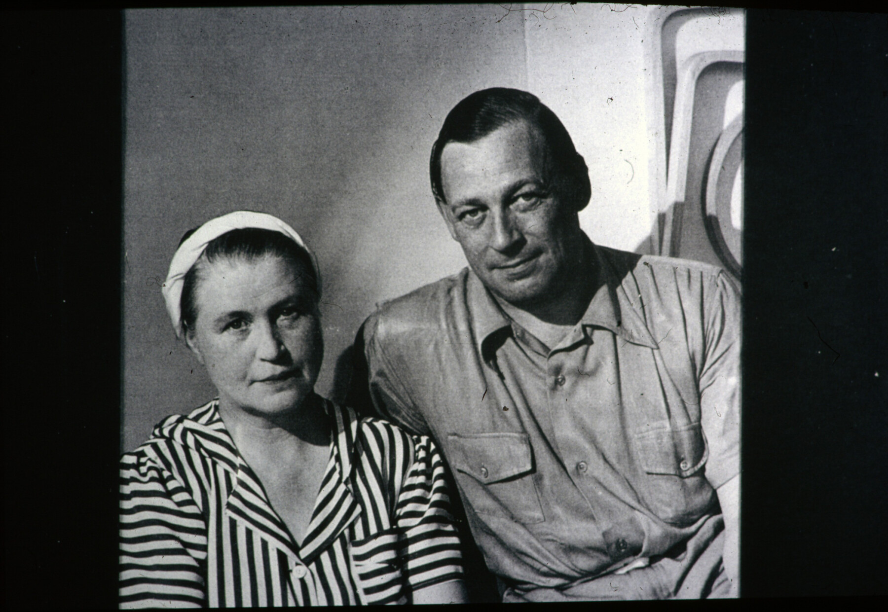 Aino Aalto, wearing a striped blouse and headscarf, sits beside Alvar Aalto in a black-and-white photograph taken indoors.