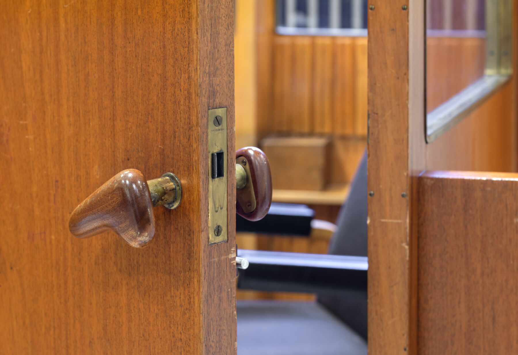 A close-up photograph shows a partially open timber door with a rounded sculptural handle, brass fittings and warm wooden surfaces of the former customer service counter at the National Pensions Institute of Finland.