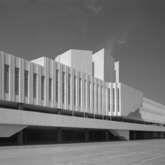 A side view of Finlandia Hall in Helsinki shows a long white façade with narrow vertical windows, geometric upper sections and a broad paved forecourt in front of the building.