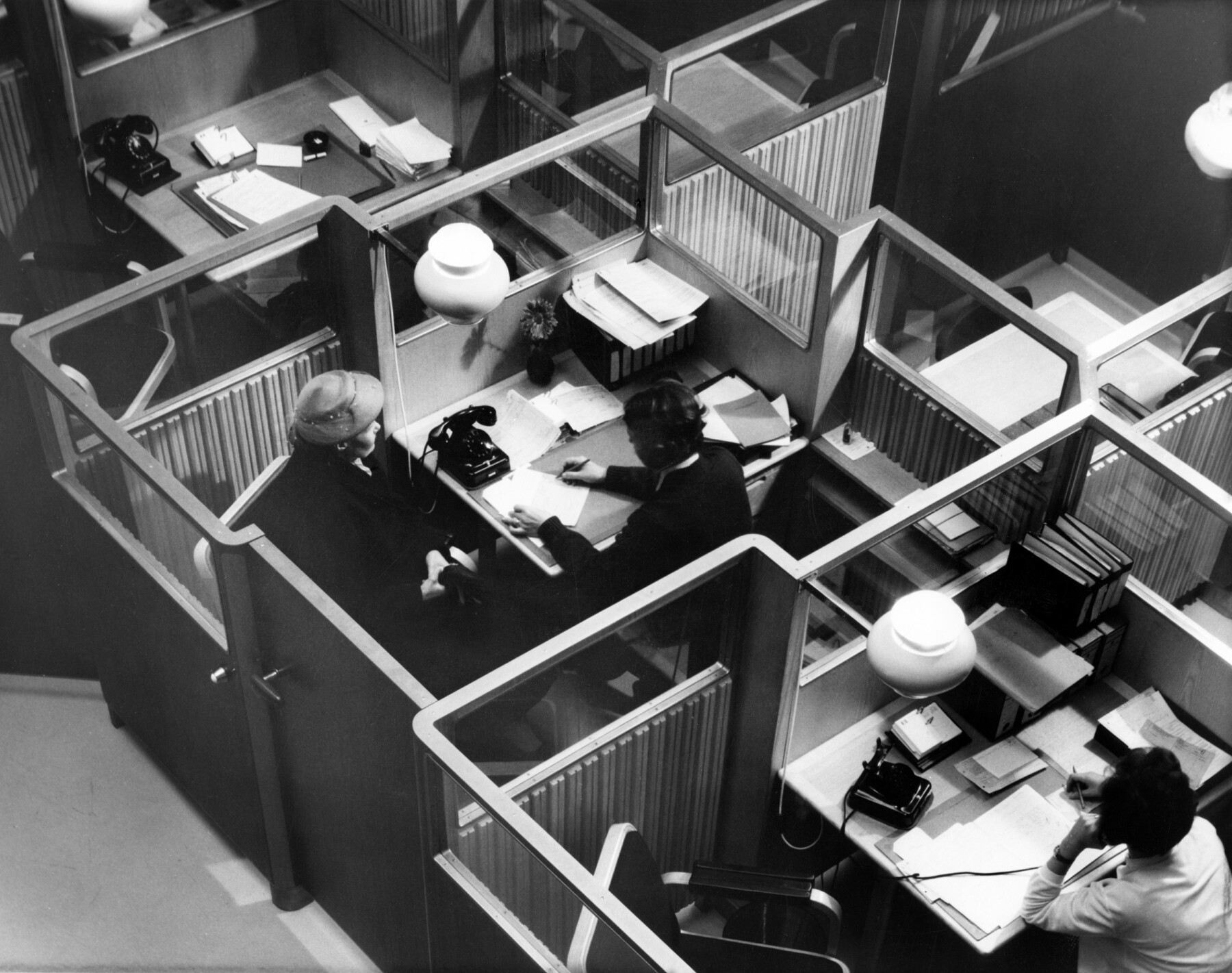 An overhead view of the National Pensions Institute (KELA) central hall in Helsinki shows Alvar Aalto’s interior design with partitioned desks, glass panels, hanging lights and office equipment from the 1950s.