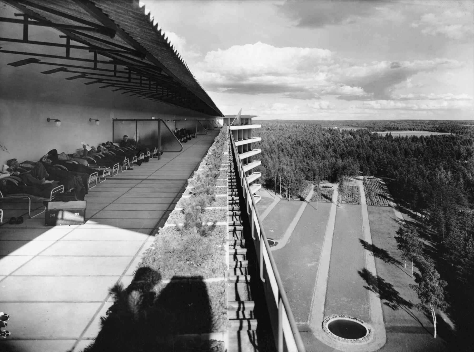 In a black-and-white photograph from 1934, patients rest on reclining chairs along the roof terrace of Paimio Sanatorium, overlooking surrounding forest and landscaped grounds.