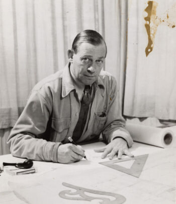 Alvar Aalto looks towards the camera while working at a desk covered with drawings, a set square and other drafting instruments.