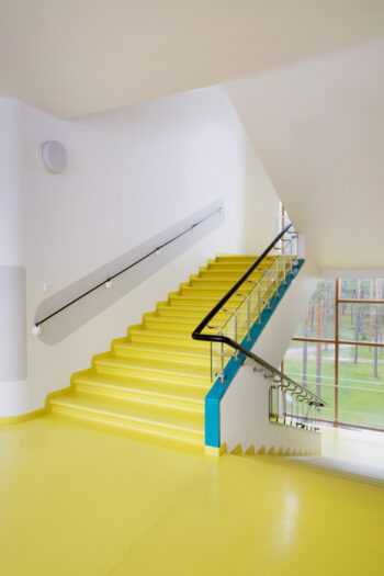A staircase in Paimio Sanatorium features vivid yellow steps and floors, black handrails, white walls and floor-to-ceiling windows facing a landscape of pine trees.