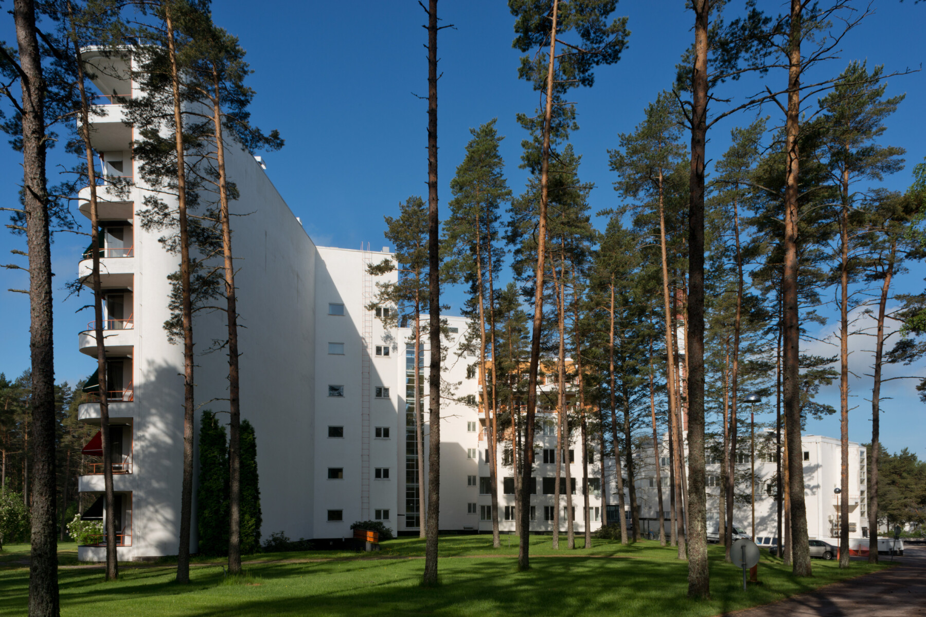 White multi-storey buildings of Paimio Sanatorium stand within a pine forest, surrounded by lawns and pathways under a clear blue sky.