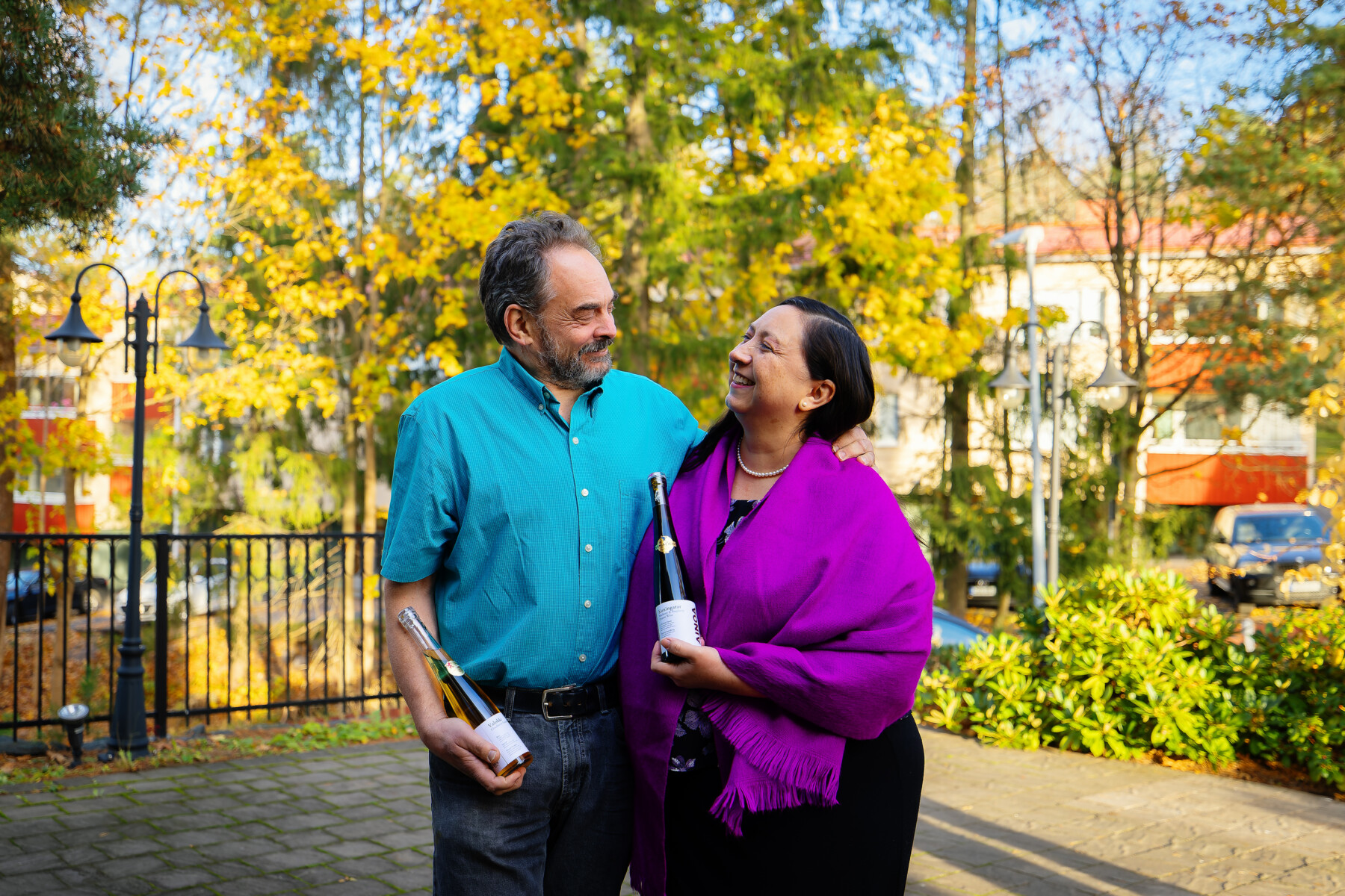 Winemakers Paola Guerrero de Cohen and David Cohen stand outdoors holding bottles of Ainoa wine, with sunlit foliage in the background.
