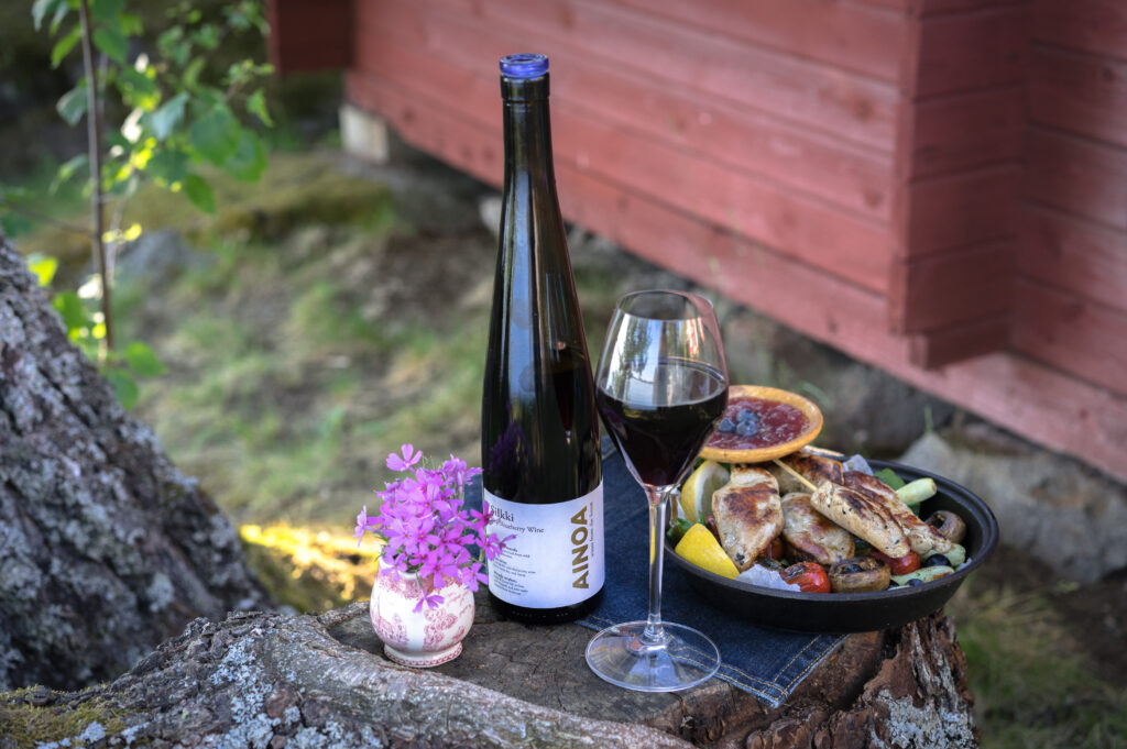 A bottle of Ainoa berry wine and a filled wine glass are displayed on a tree stump beside a plate of grilled food, with a red wooden building in the background.