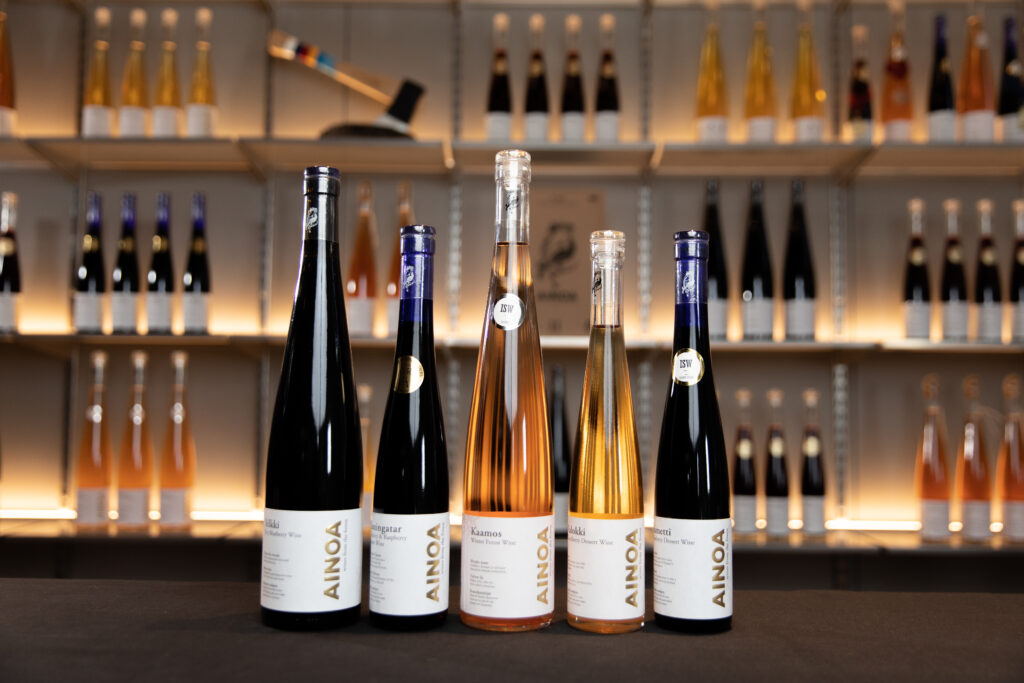 Five bottles from Ainoa Winery’s range of berry wines and dessert wines stand on a counter, with shelves of illuminated bottles blurred in the background.