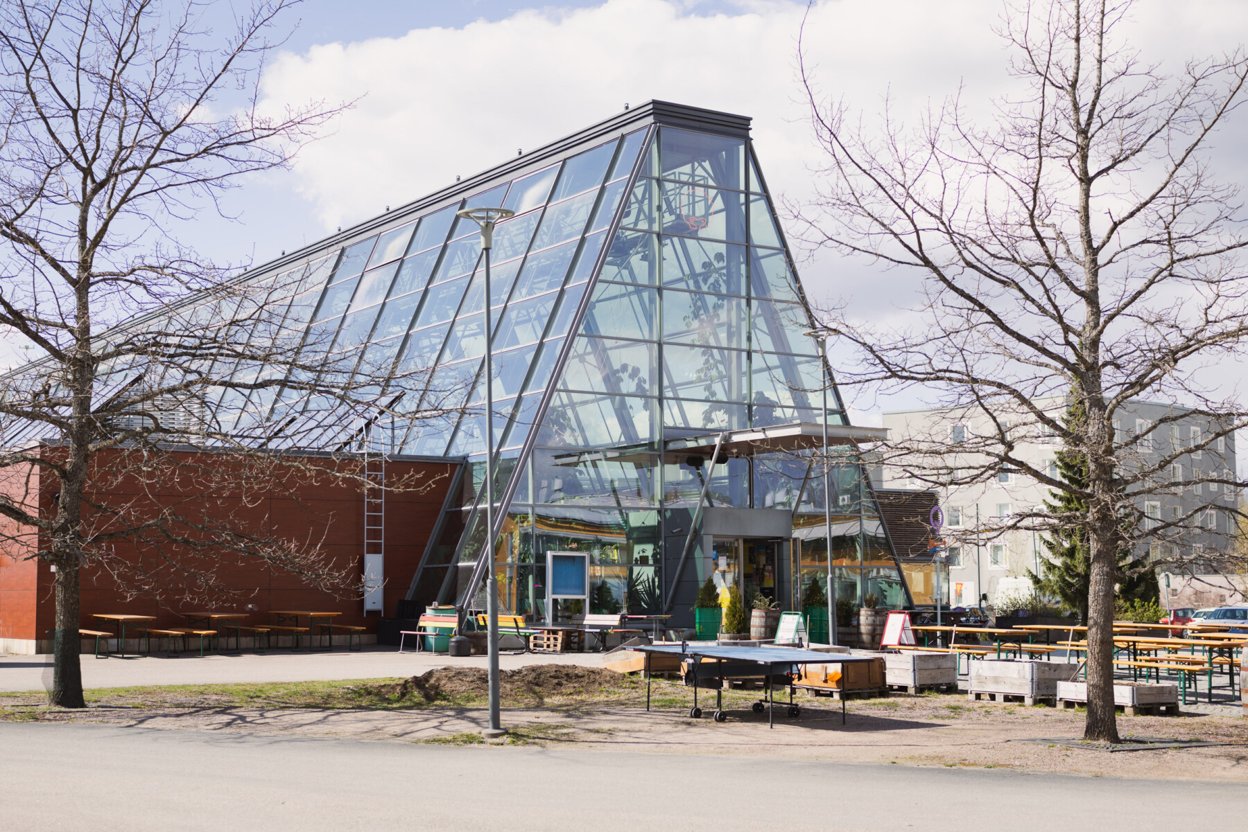 A glass-fronted greenhouse building in Viikki houses CoolHead Brew’s brewery and taproom, with outdoor seating arranged around the entrance.