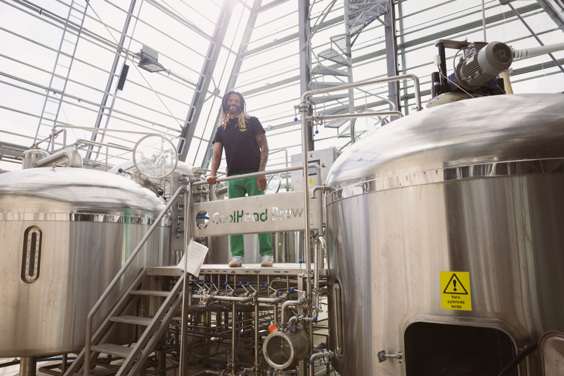 CoolHead founder Cleber Gonçalves stands on a raised metal platform between large stainless-steel brewing tanks inside a bright industrial brewery.
