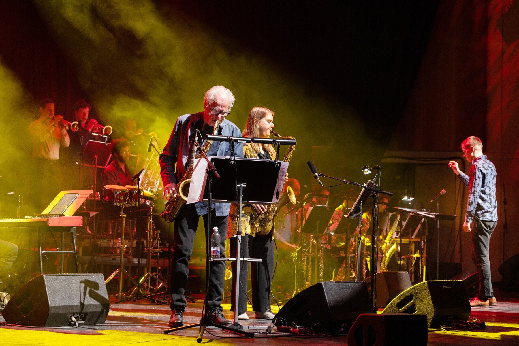 Two saxophonists, one man and one woman, play in front of a group of other musicians on a stage.