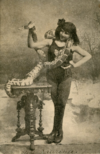Circus performer Elvira Bono poses with a large snake draped around her shoulders beside an ornate table in an early 20th-century black-and-white photograph.