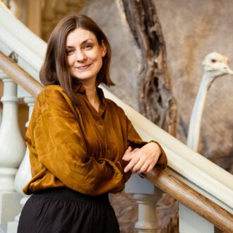 Author Iida Turpeinen leans on a staircase railing inside the Finnish Museum of Natural History, with part of a large bird exhibit visible behind her.