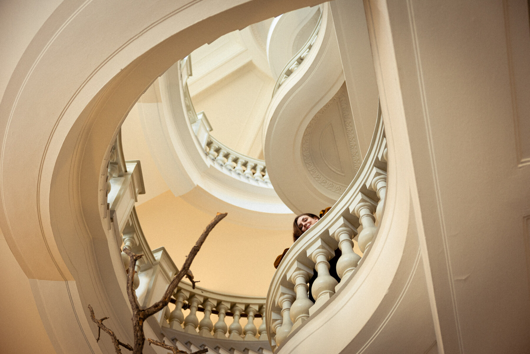 Author Iida Turpeinen looks down from a curved staircase inside the Finnish Museum of Natural History, framed by white balustrades and arches.