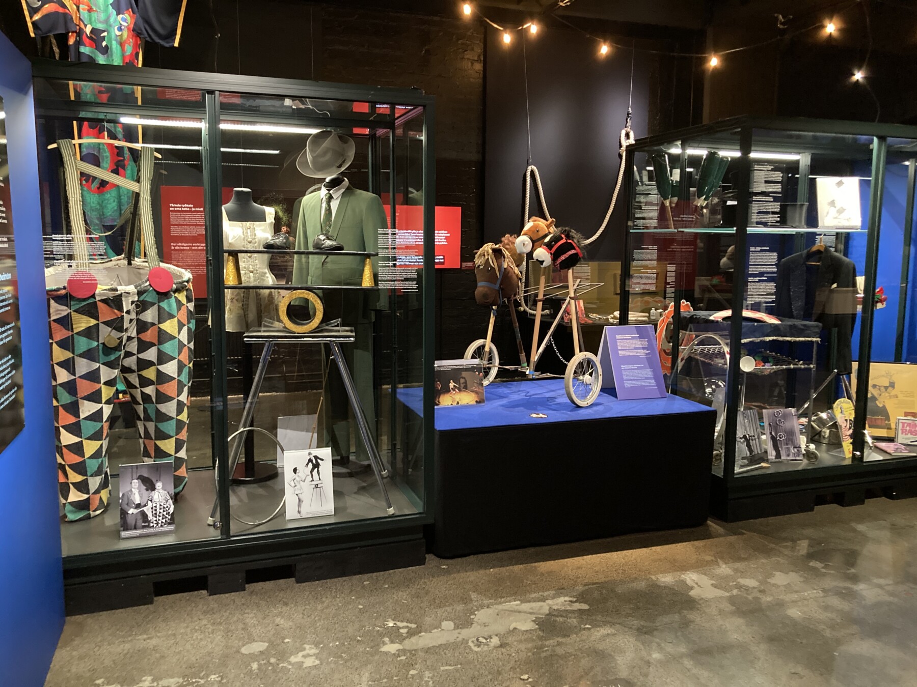 Circus costumes, props and performance equipment are displayed in glass cases, including colourful clown trousers, a suit and hat, a circus ring and three hobby horses on wheels.