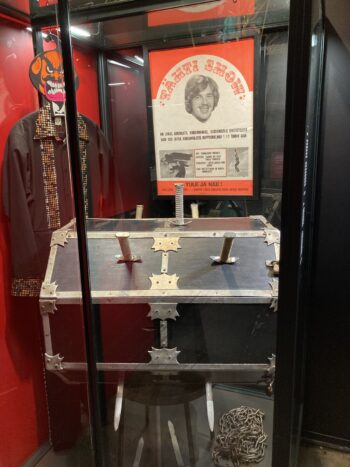 A black trunk with swords thrust through it sits inside a museum exhibit about Finnish circus performer Aimo Leikas, alongside chains, a costume and a historic show poster.
