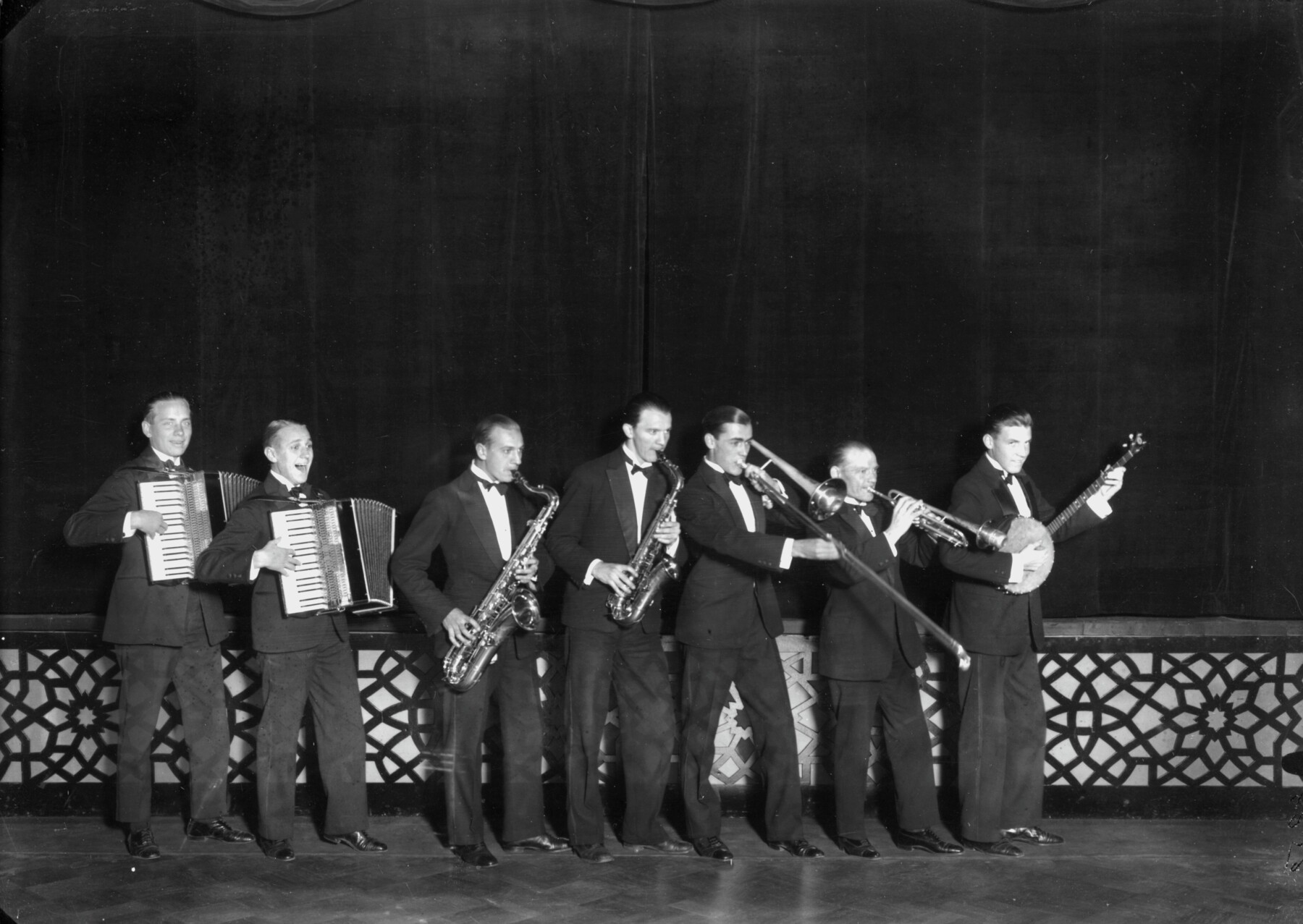 Seven musicians in suits pose in a row, including two accordionists, two saxophonists, a trombonist, a trumpeter and a banjo player.
