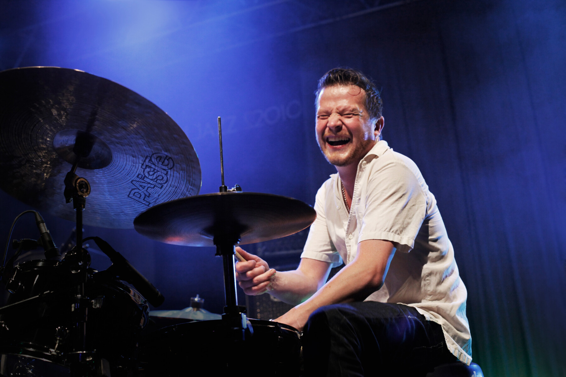 A man with closed eyes smiles as he plays a drum set.