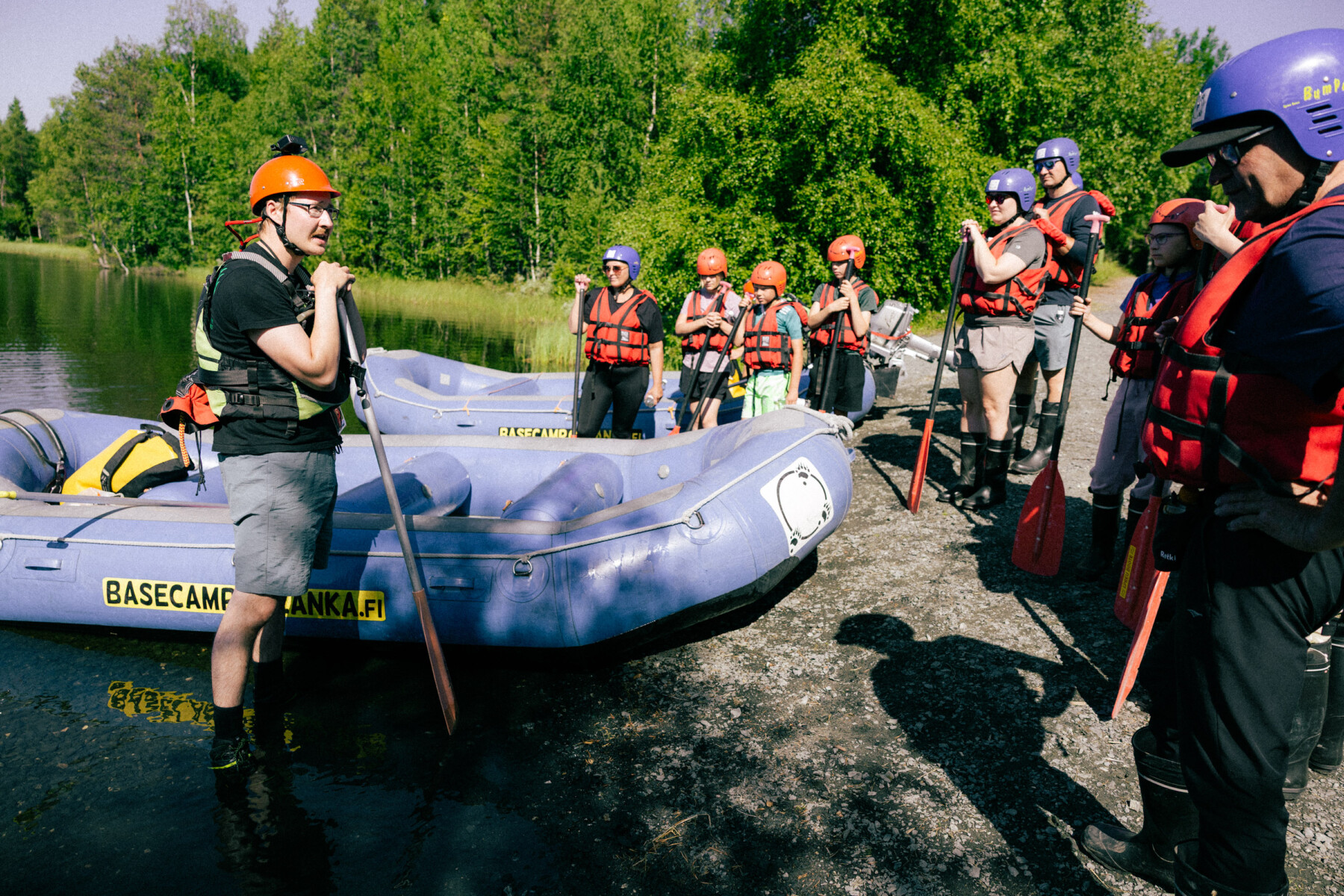 A rafting guide wearing an orange helmet speaks to a group of adults and children gathered beside inflatable boats on a riverbank in northern Finland.