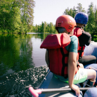 Two people wearing helmets and red life jackets sit at the front of an inflatable raft drifting along a calm forest river in northern Finland.