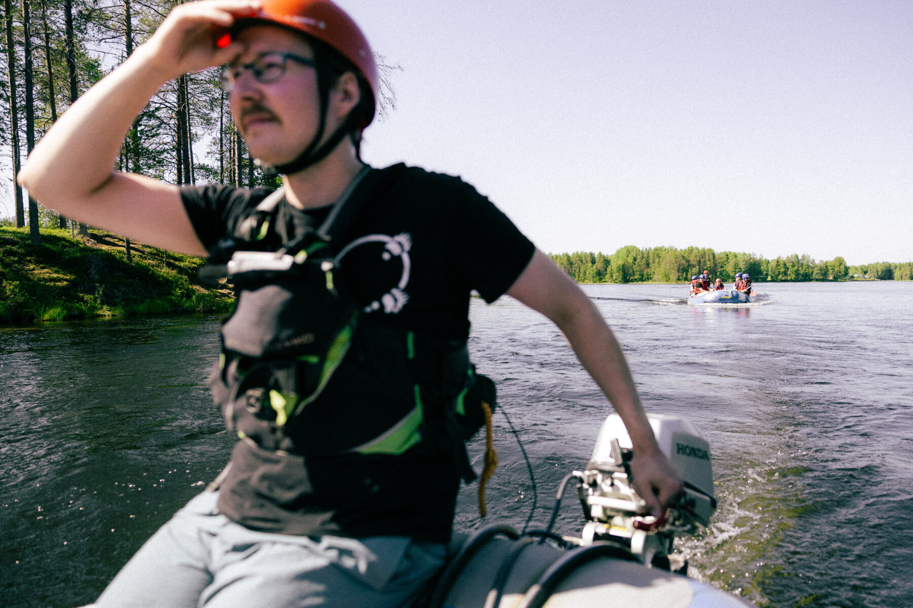A guide sitting at the back of an inflatable raft shields his eyes from the bright summer light while using the outboard motor to steer across calm water bordered by forest.