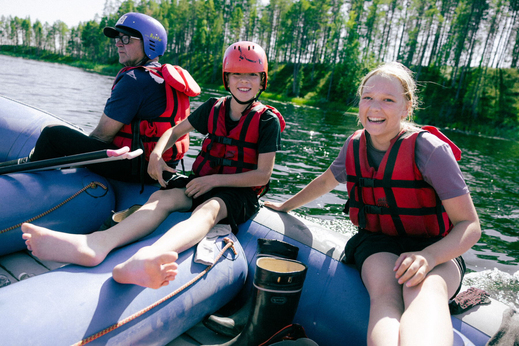 Three people wearing red life jackets sit in an inflatable raft on a river, with pine trees lining the shore behind them.