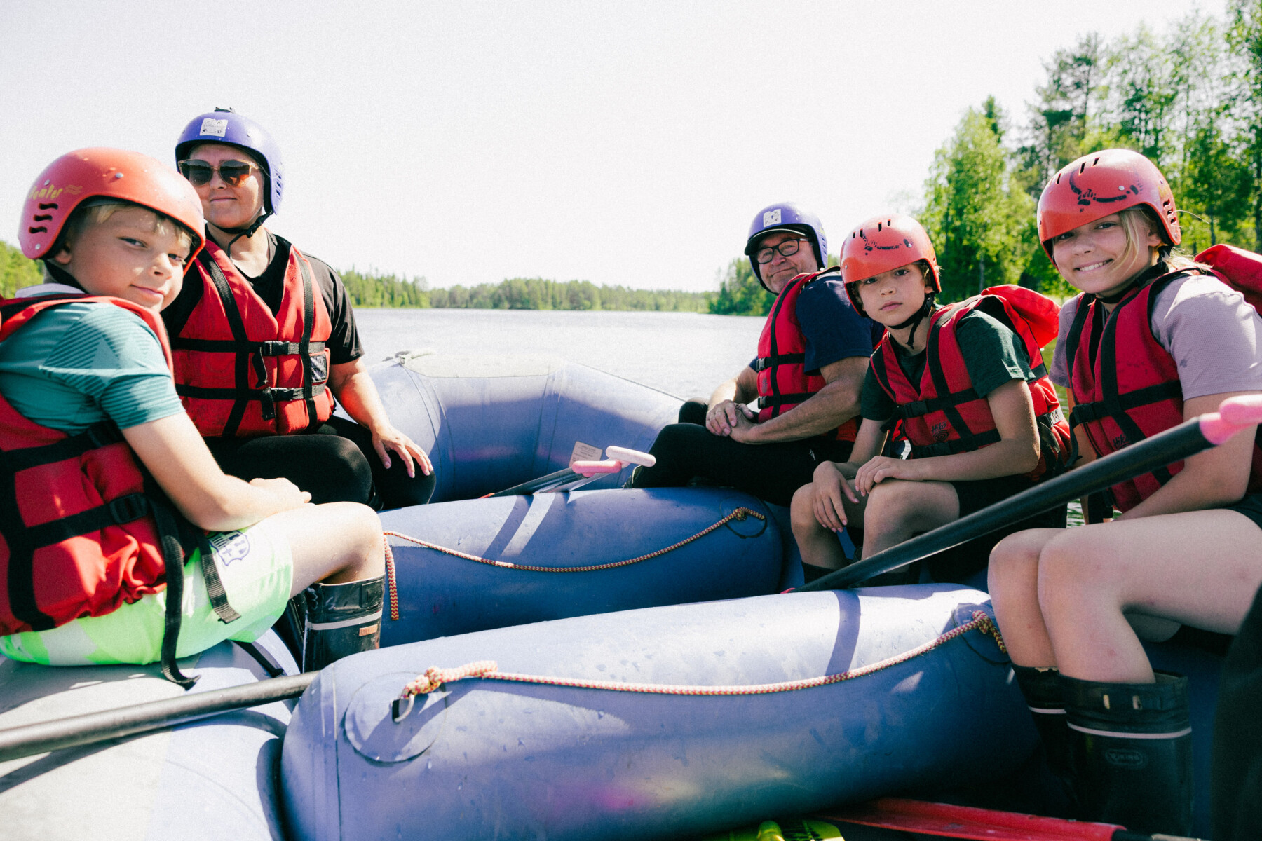 Two inflatable rafts carrying people in safety gear float on a river under bright daylight in northern Finland.
