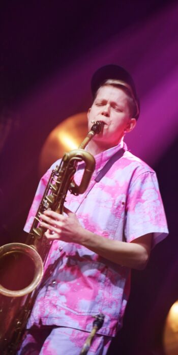 A woman in a baseball cap plays a baritone saxophone, with colourful stage lighting in the background.
