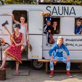 Seven female circus performers pose in and around a white caravan labelled “SAUNA,” surrounded by objects associated with Finnish life, including firewood, an axe, a violin and skis.
