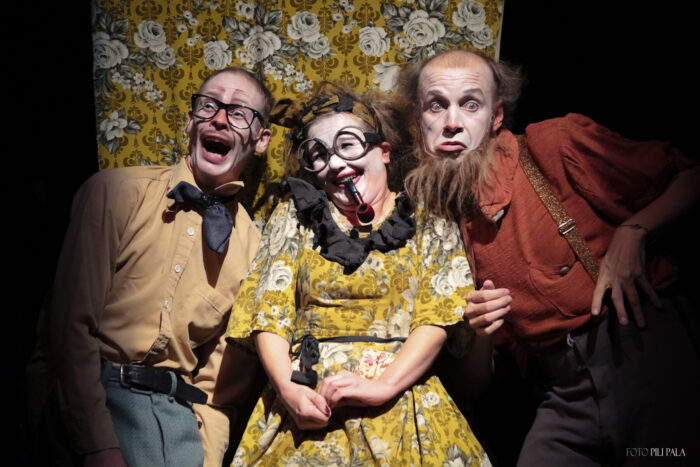 Three performers in colourful costumes and theatrical makeup pose together with exaggerated expressions against a floral backdrop during Kallo Collective’s Members of Our Limbs.