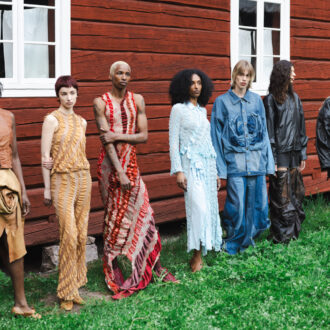 Seven models stand on the lawn beside a red wooden building, wearing garments that range from textured dresses to oversized denim and black outerwear.