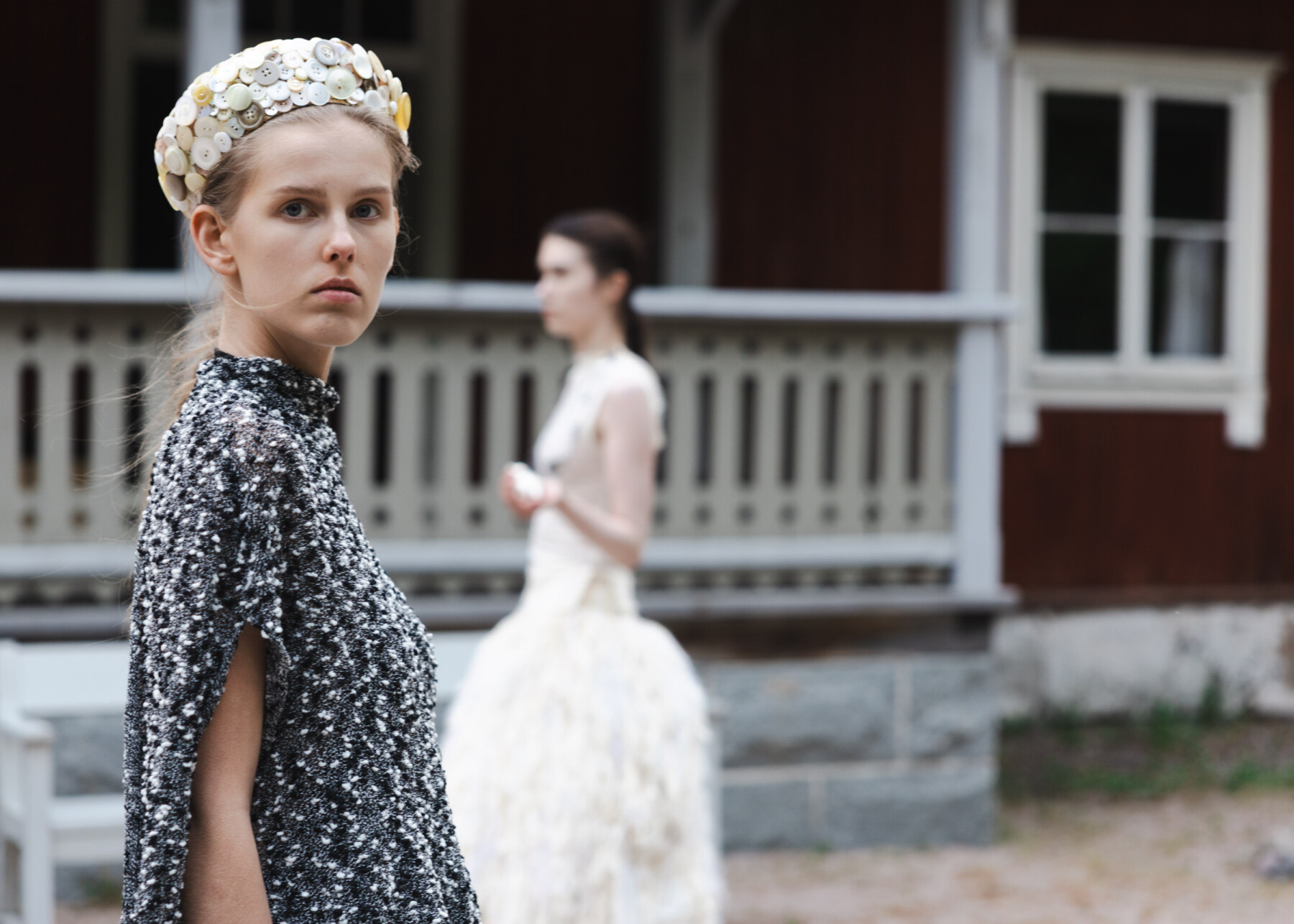 A model wearing a textured black-and-white garment and a headpiece made from recycled materials stands in front of a traditional wooden building, while another model appears blurred in the background.