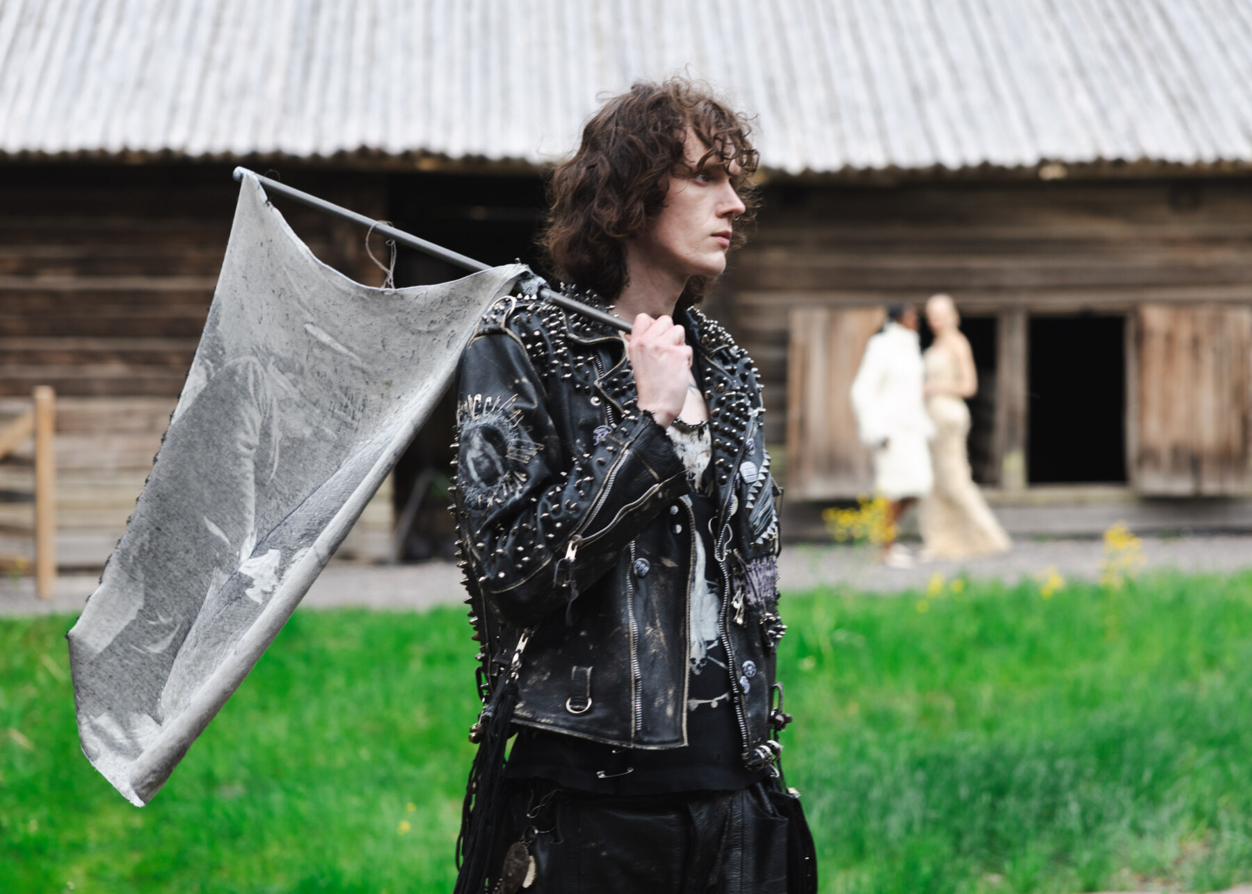 A model wearing a studded black leather outfit carries a grey flag across a grassy field, with other models blurred in the background near a traditional Finnish wooden building.