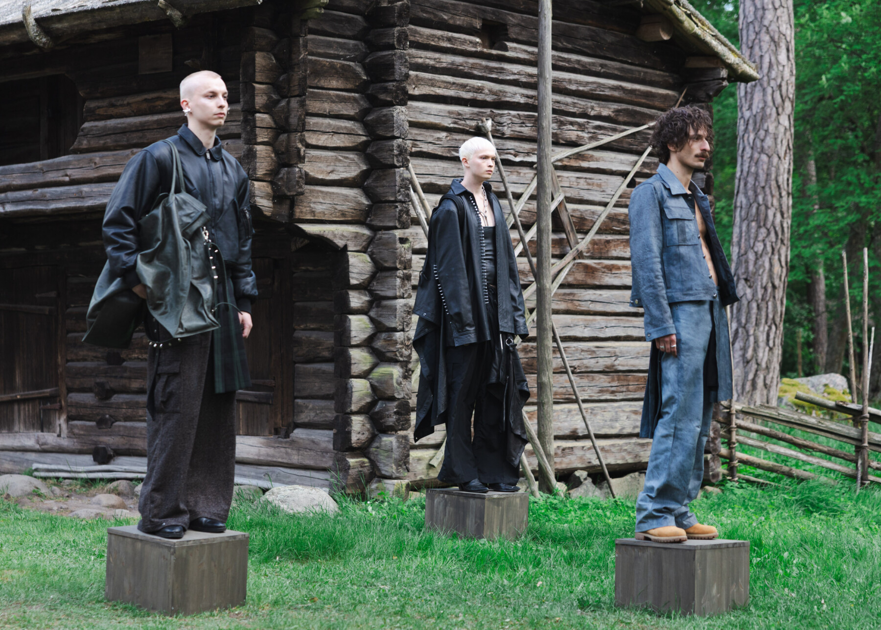 Three models stand on wooden plinths in the courtyard of a historic log building, wearing dark layered garments and denim pieces from Jim Bergström’s collection.