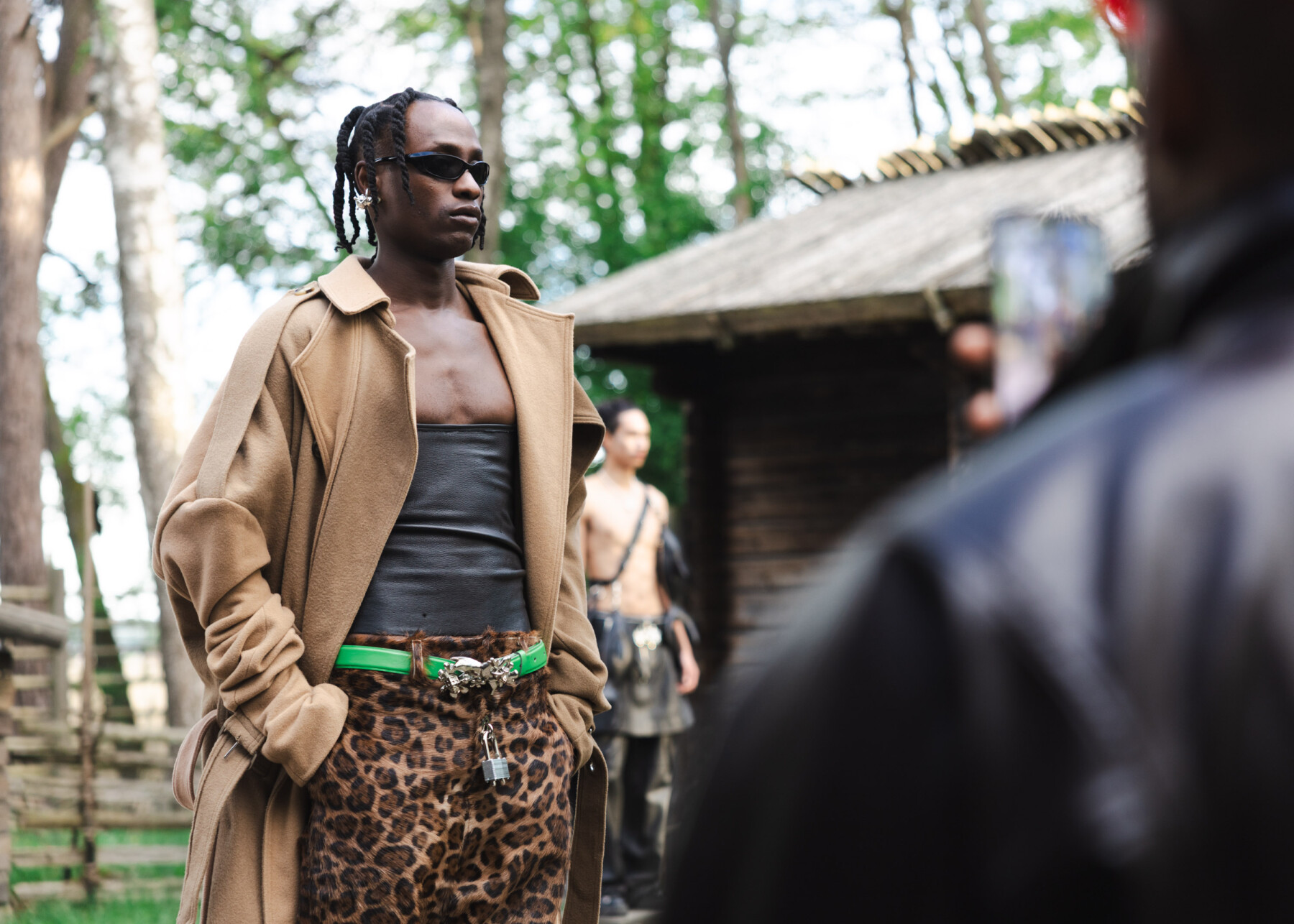 A model wearing a long tan coat, a dark top, leopard-print trousers and green belt poses outdoors at a fashion show in Seurasaari, Helsinki.