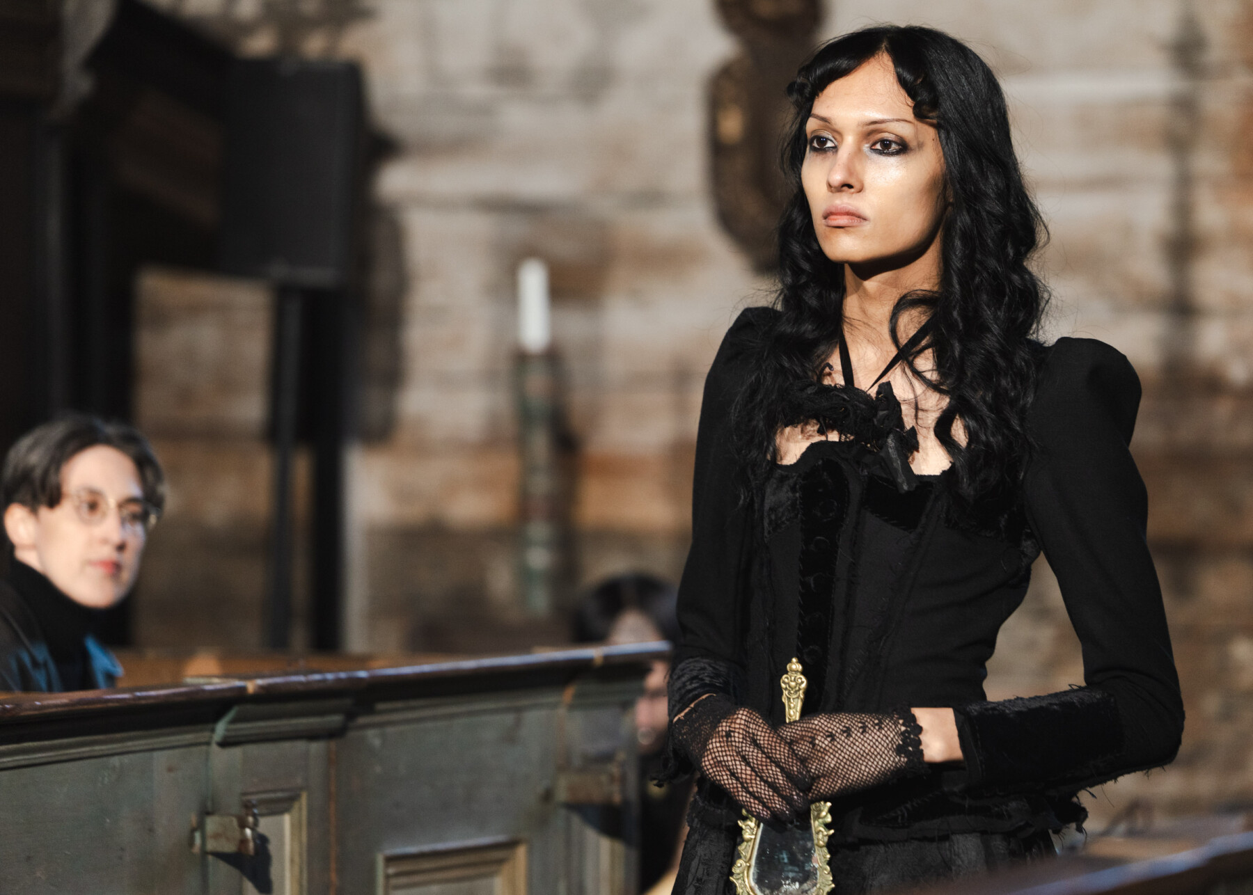 A model wearing a fitted black dress and mesh gloves holds an antique mirror during Linda Kokkonen’s runway show in Karuna Church, as audience members watch from wooden pews.