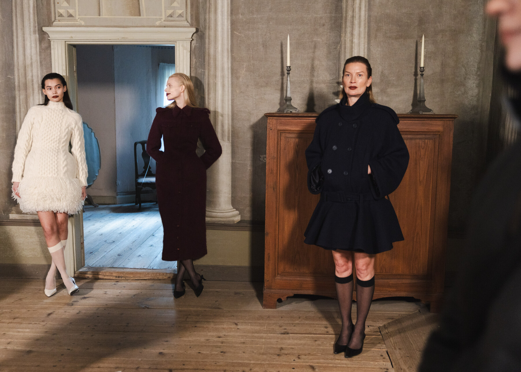 Three models wearing white and black designs by Idaliina Friman stand in a historic interior on the island of Seurasaari during a Helsinki Fashion Week event.