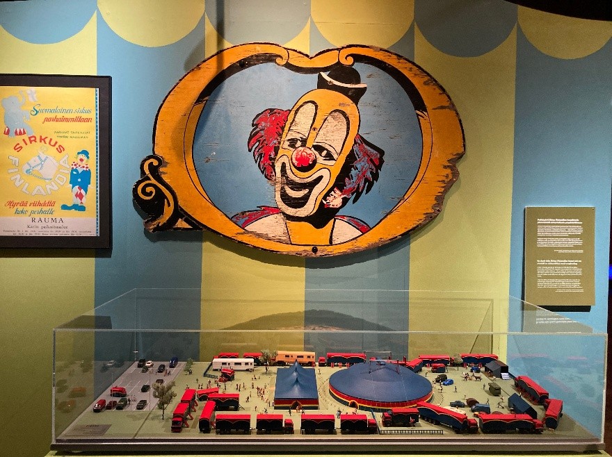 A large vintage clown sign mounted on a striped exhibition wall hangs above a detailed scale model of a travelling circus site with tents, vehicles and crowds of miniature visitors.
