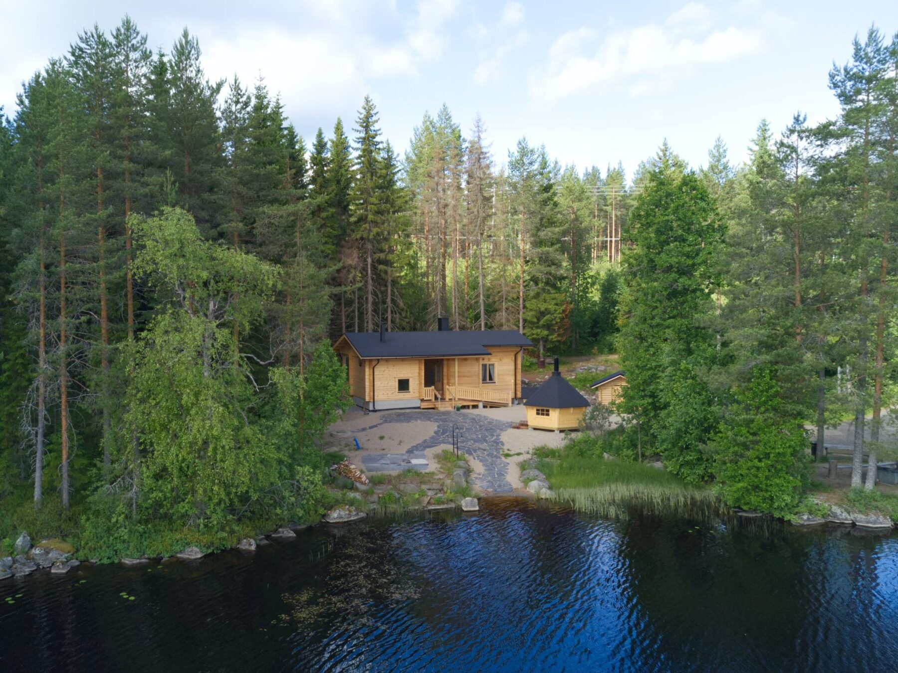 A wooden sauna and outbuilding are on a lake shore in a forest.