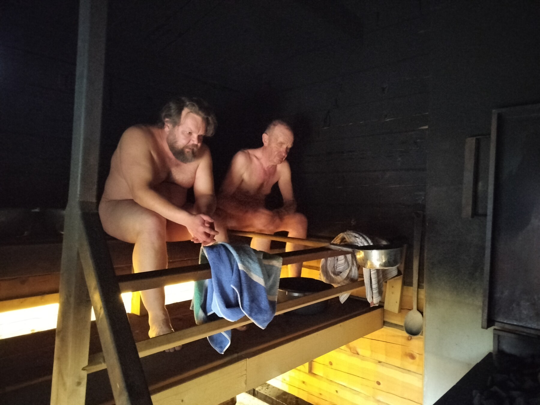 Two men sit in a dark sauna. One throws water onto the stove.