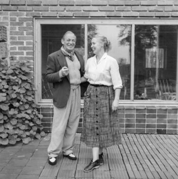 Alvar Aalto, wearing a dark jacket, light jumper and loose trousers, stands beside Elissa Aalto, who wears a light blouse and patterned skirt, outside their summer house in Muuratsalo, Finland.