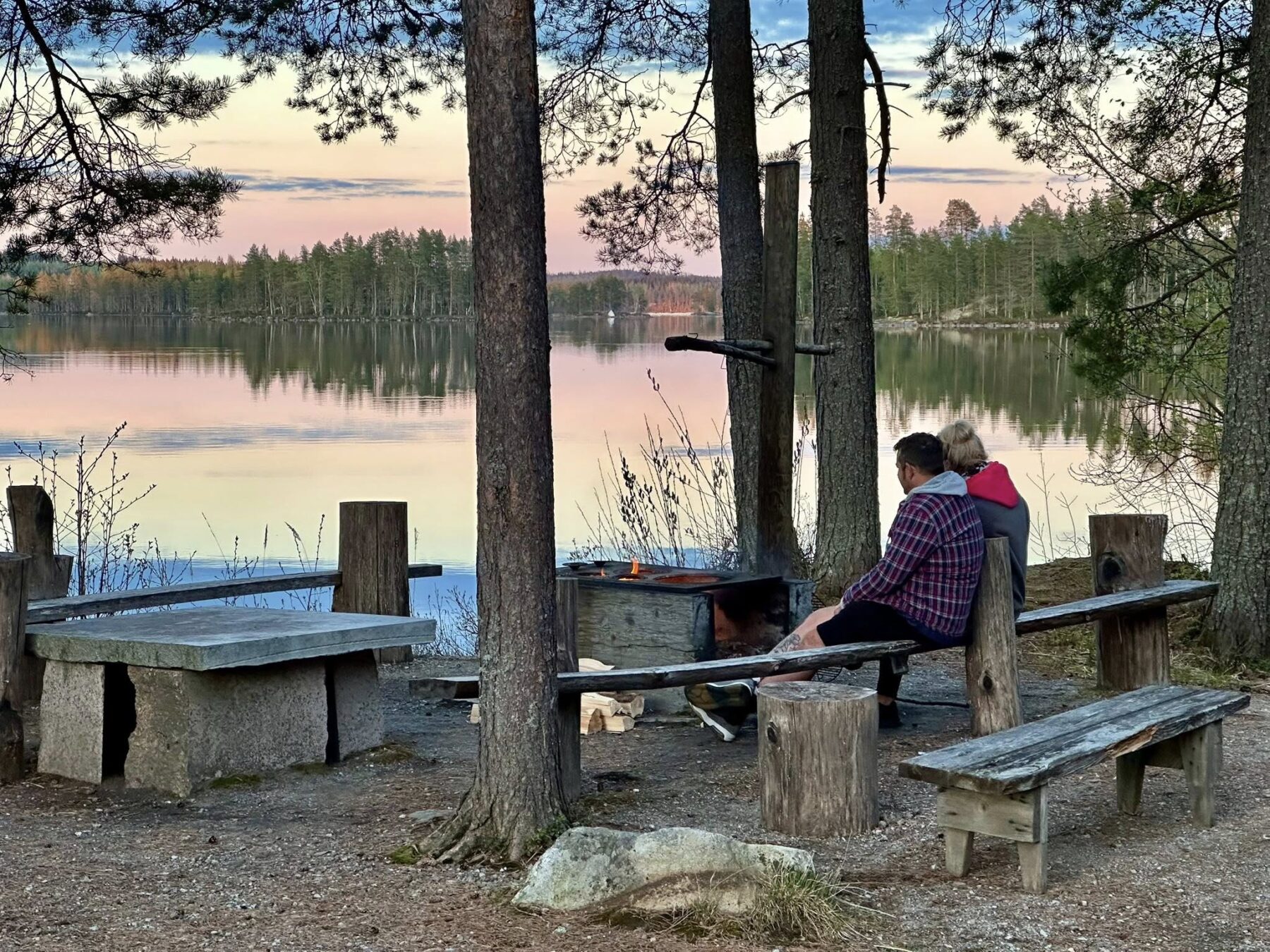 Two people sit on a wooden bench next to a fire in a wooded area. A lake is in the background.