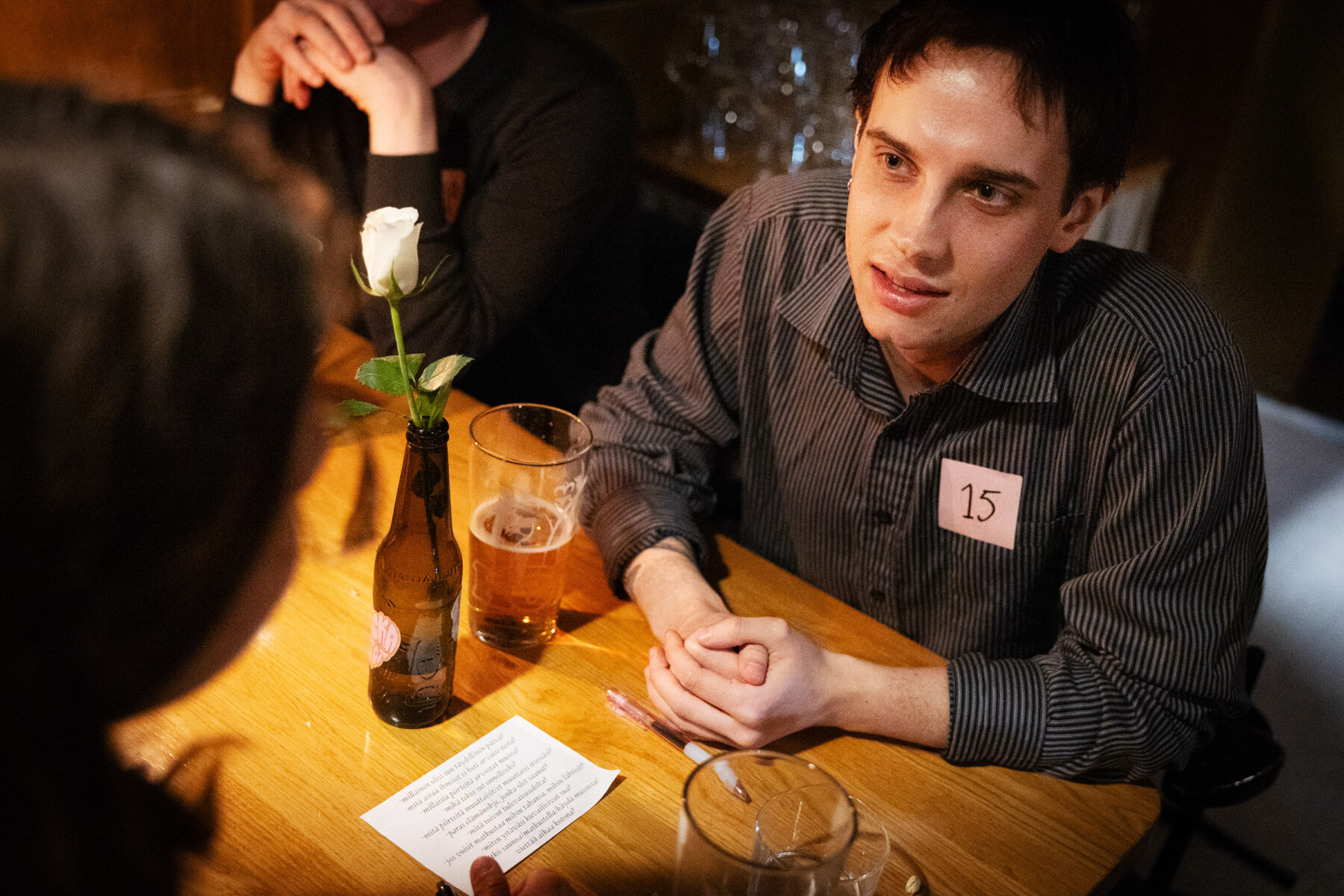 Com um adesivo numerado na camisa, um homem conversa atentamente com sua parceira de mesa. Entre os dois, há um copo de cerveja, um cartão de instruções e uma delicada flor branca.