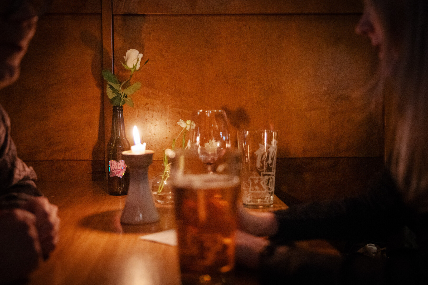 Bebidas, uma vela acesa e um arranjo floral compõem a aconchegante mesa de um bar, tornando o cenário perfeito para um encontro marcado pela conversa.