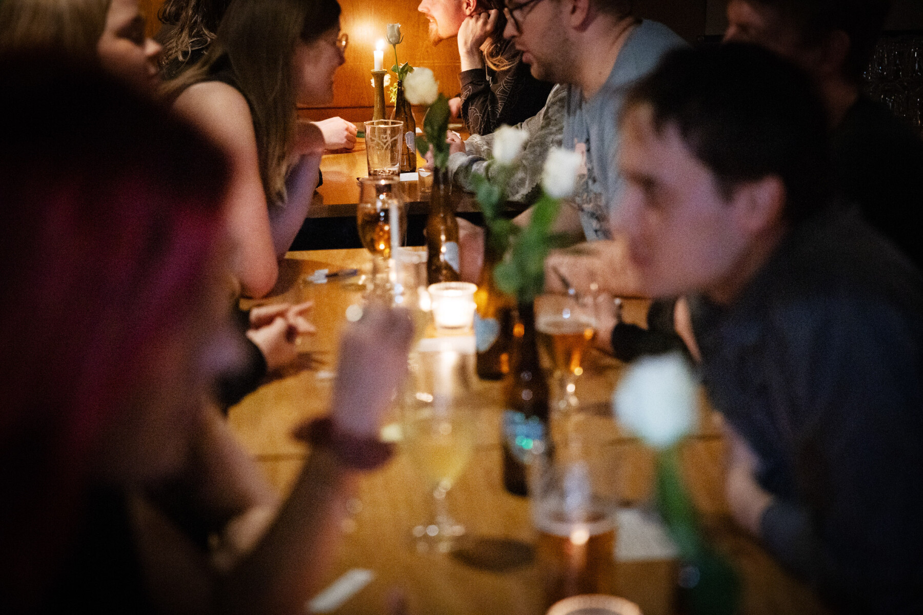 Participantes sentados frente a frente, formam pares ao longo de uma grande mesa de madeira em um bar iluminado por luz de velas. Inclinados para a frente, conversam enquanto compartilham bebidas e o clima intimista do evento.