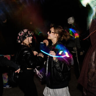 Duas pessoas dançam e conversam sob feixes de luz colorida em uma escura rave underground em Helsinque, enquanto outros participantes se reúnem ao redor delas nas sombras.