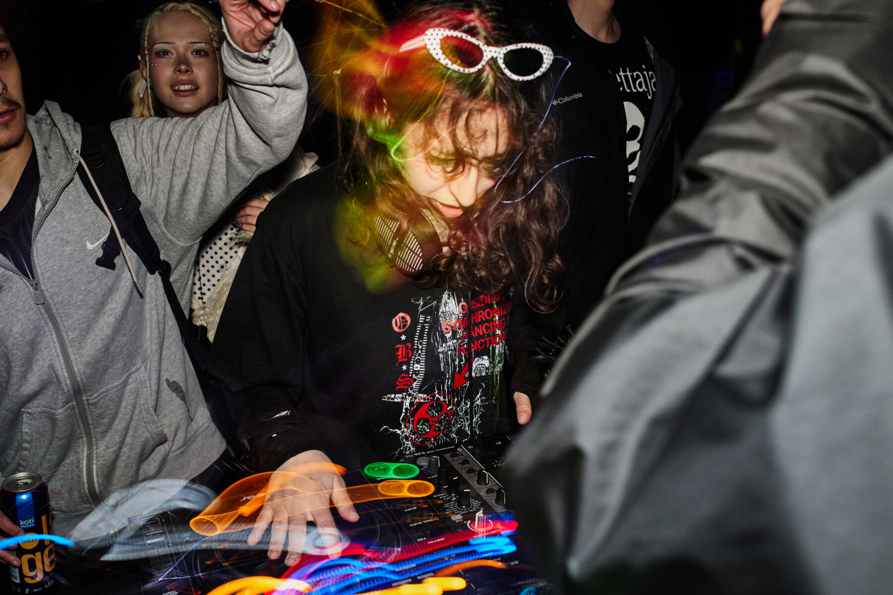Uma DJ usando óculos escuros com armação branca mixa músicas em um controlador, enquanto rastros coloridos de luz atravessam uma pista de dança lotada em uma rave underground.