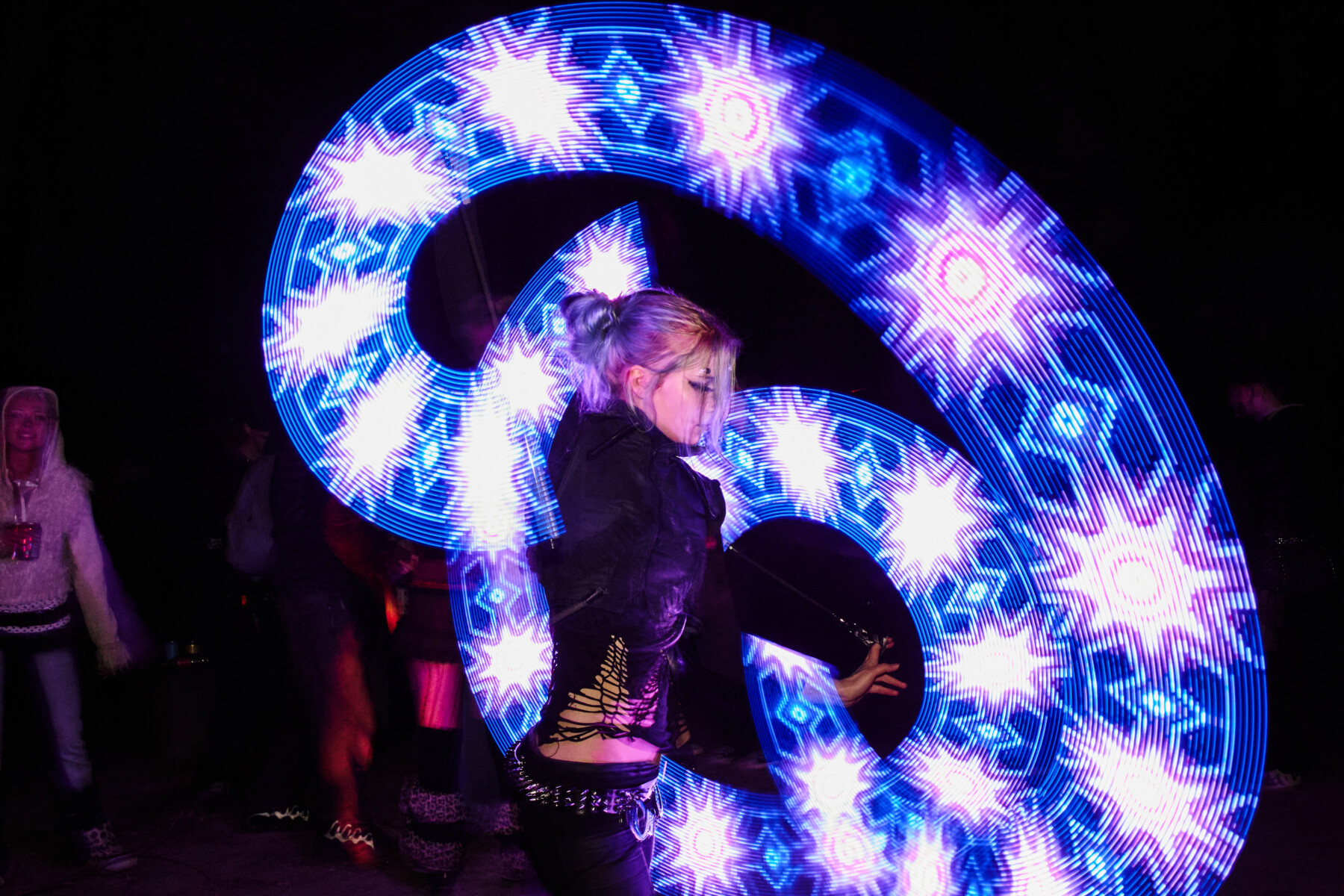 Uma mulher se apresenta com acessórios luminosos de LED criando padrões azuis e brancos brilhantes, girando pela escuridão, durante uma rave ao ar livre em Helsinque.